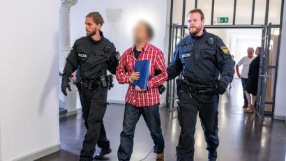 Bild: Armin Weigel
Der Angeklagte, der dreifach gefesselt ist, wird von Polizisten zum Gerichtssaal geführt.