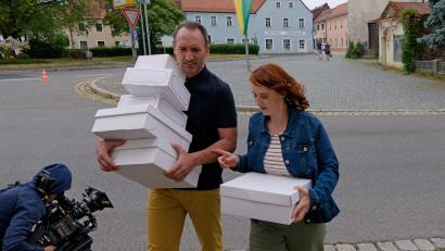 Bild: Norbert Grüner/Set- und Standbildfotograf Stonewoold Film e. V. 
Drehstart Montag, 8.15 Uhr, Klappe 1: Die Schauspielerin und Produzentin Christina Baumer in der Hauptrolle als Magdalena Brandt und Schauspieler Jürgen Fischer als Sepp Hofmann tragen Kuchenschachteln über den Deschplatz in Tirschenreuth.