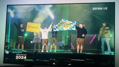 Bild: Sutrich/exb
Mit einem Foto vom heimischen Sofa aus wurde der Auftritt des Sängers Tream (Dritter von rechts, leicht verdeckt) aus dem Schwandorfer Ortsteil Büchelkühn vom Fernsehbild weg dokumentiert. Tream war Teil der großen ZDF-Silvestershow 2023 in Berlin. Seine Mitstreiter halten ein Büchelkühn-Ortsschild in die Höhe.