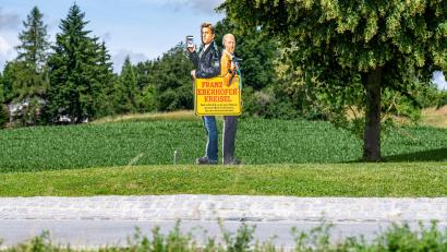 Archivbild: Armin Weigel/dpa
Ein Bild mit den Darstellern der Krimireihe um den Dorfpolizisten Franz Eberhofer und Co-Ermittler Rudi steht auf dem Franz-Eberhofer-Kreisel. Nun wurde die Linde von Unbekannten gefällt.