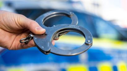 Bild: David Inderlied/dpa/Illustration
Eine Hand hält Handschellen vor einen Streifenwagen der Polizei.