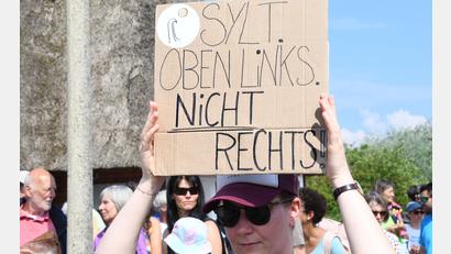 Symbolbild: Lea Sarah Albert/dpa
„Sylt oben links – nicht rechts“ steht auf einem Plakat, das eine Frau bei einer Mahnwache auf Sylt in der Hand hält.