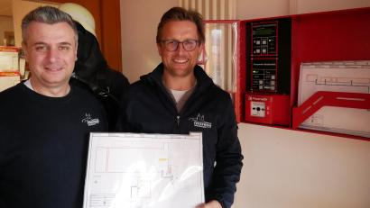 Bild: flm
Florian Meyer (links) und Kommandant Tobias Tippmann vor der sogenannten "Brandmeldezentrale" im Gerätehaus. Dies ist im Brandfall der erste Anlaufpunkt für die Feuerwehr. Hier lagern auch die Laufkarten, die zu den ausgelösten Meldern führen.