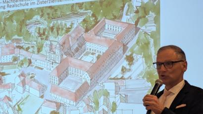 Bild: pz
Bürgermeister Bernd Sommer während seines Vortrags in der jüngsten Bürgerversammlung. Auf der Leinwand zu sehen ist eine Skizze mit einer Ansicht des Klosters, an dem der Anbau (oberer Bildrand) für die erweiterte Realschule bereits verwirklicht ist.