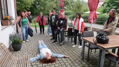 Bild: bsc
Zur Freude der Teilnehmer wurden bei der Drehort-Tour durch Friedenfels auch Szenen nachgespielt, wie hier von Ulla Britta Baumer (auf dem Boden liegend) die Szenerie, als sie im Film mausetot vor der Eingangstür des Friedenfelser Dorfladens liegen musste.