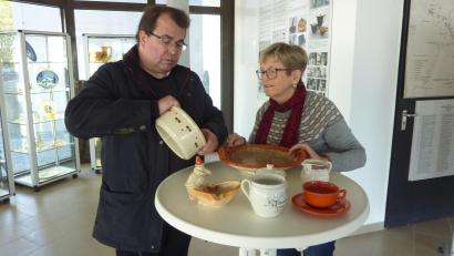 Bild: Archiv Helga Katrin Stano
Pfarrer Haußmann, der ein Kenner der Schwarzenfelder Tonwaren ist und auch schon darüber referierte, mit Helga Katrin Stano, deren Mutter noch in der Heferlbude arbeitete. Sie trug mit Unterstützung von Herrn Linzmeier eine stattliche Sammlung von Steingutstücken aus der Tonwarenfabrik zusammen, die man im Eingangsbereich des Schwarzenfelder Rathaus sehen kann. In Pfarrer Haußmanns Hand eine Schüssel (Alltagsgeschirr) mit Schablonendekor. Die Tasse im Vordergrund ist das älteste Exponat und dürfte von 1905 sein. Der Krug rechts ist aus den 20er-30er Jahren. Tasse mit Untertasse ist minderwertige Kriegsware. Der Obstteller und die Nippesfigur haben Laufglasur.
