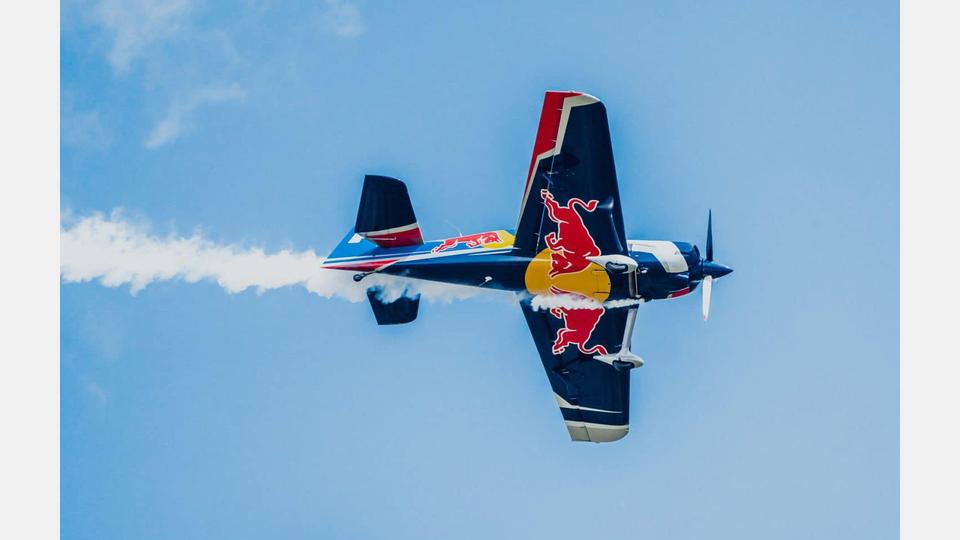 Ein Kunstflugzeug in den Farben von Red Bull (Blau, Weiß, Rot und Gelb) fliegt kopfüber am klaren blauen Himmel und hinterlässt eine weiße Rauchspur. Bild: Ultralight Club Cheb