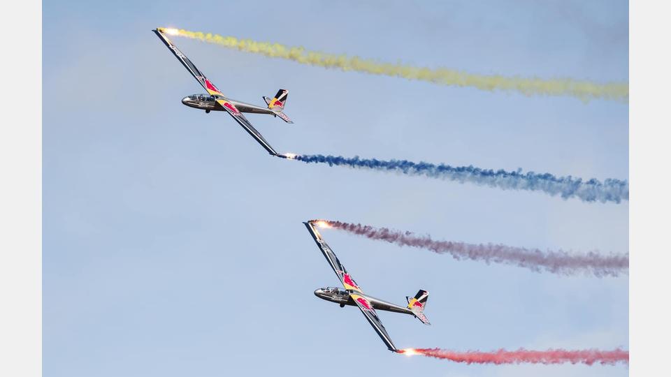 Zwei Kunstflugzeuge in den Farben von Red Bull (Blau, Weiß, Rot und Gelb) fliegen nebeneinander am blauen Himmel. Sie hinterlassen vier farbige Rauchspuren in Gelb, Blau, Lila und Rot. Bild: Ultralight Club Cheb